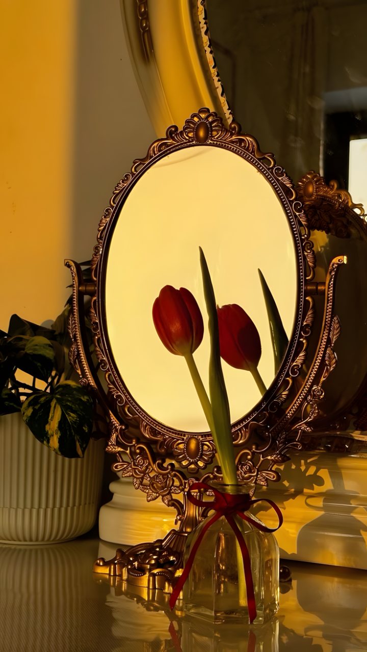 An ornate antique mirror reflects two red tulips beside a glass vase on a warmly lit table.