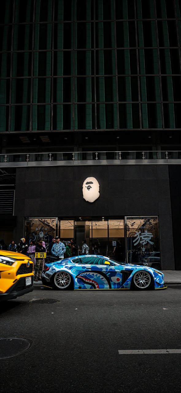 Blue sports car with a shark camouflage wrap parked outside a modern dark building, with a yellow vehicle and people nearby.