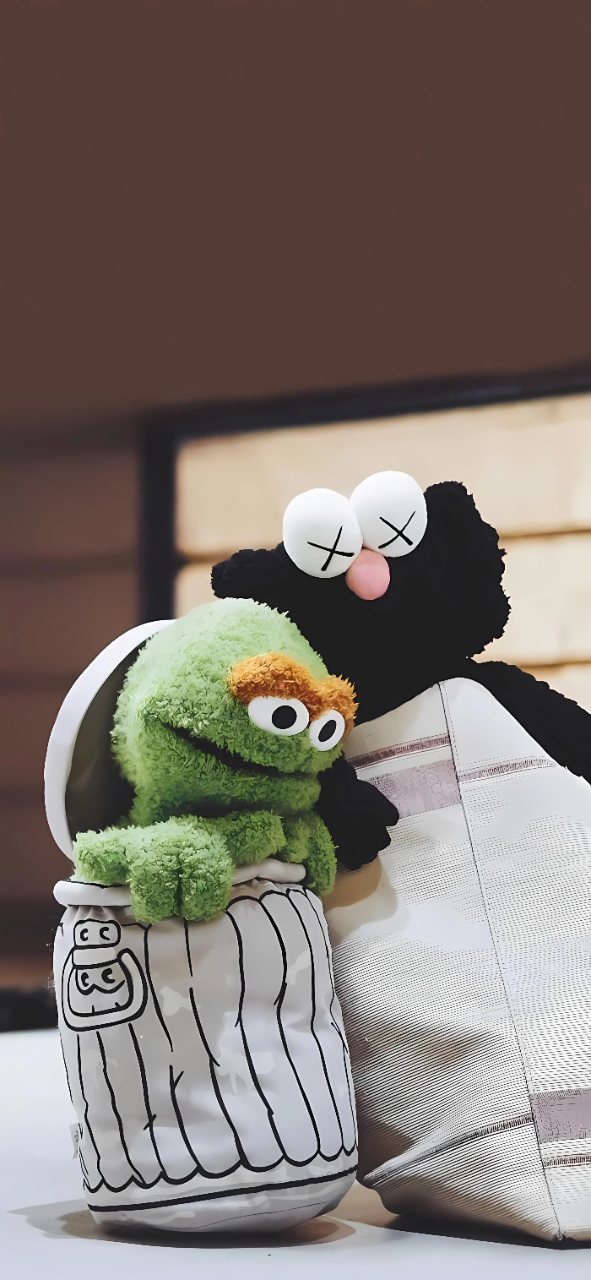 Cookie Monster and Elmo plush toys cuddling together beside a black plush character in a softly lit room.