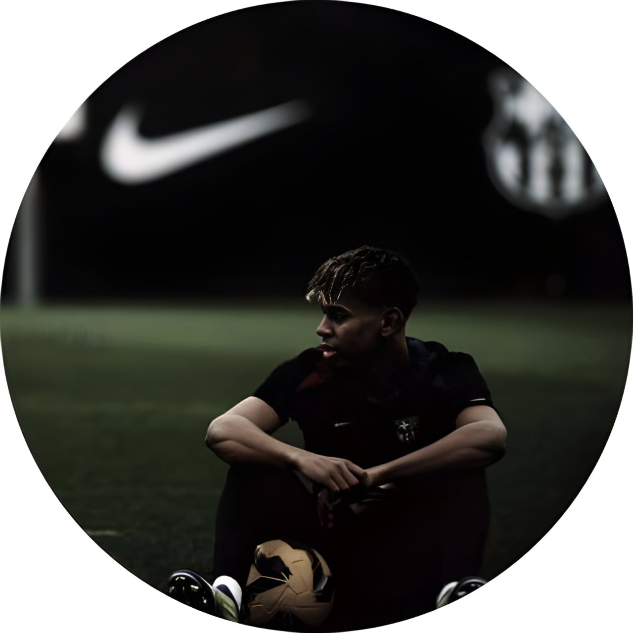 A young football player sits on a field at night with a soccer ball between his feet, surrounded by dark shadows and blurred stadium lights.