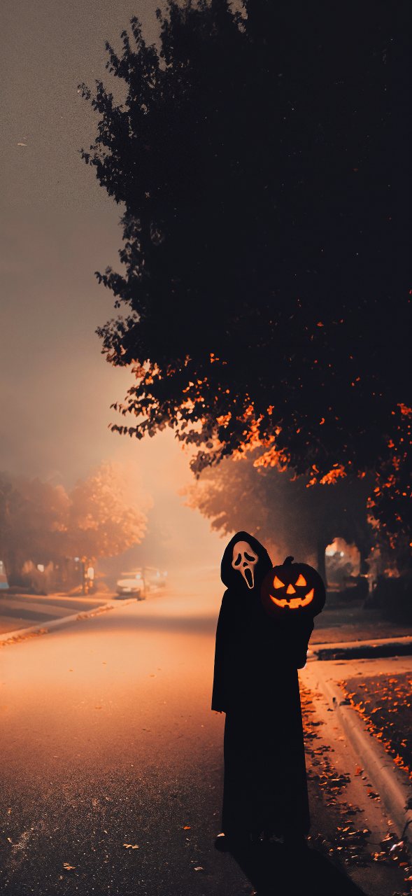 Ghostface stands on a dim autumn street holding a glowing jack-o'-lantern beneath dark trees at dusk.