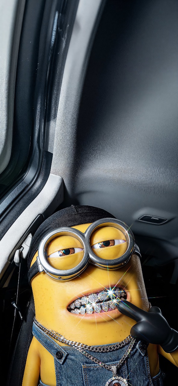 A yellow Minion wearing goggles and denim overalls sits inside a car, showing a sparkling diamond grill while a gloved hand points toward its mouth.