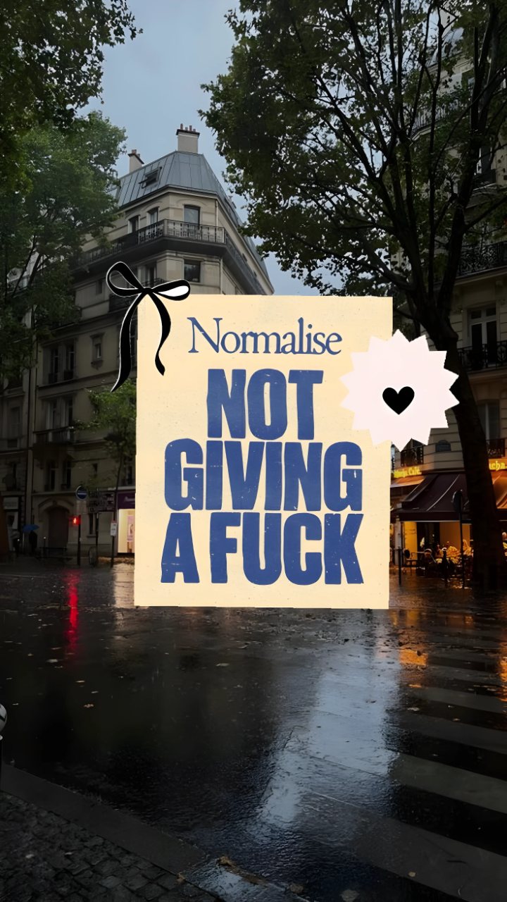 A rainy city street with a large sign reading “Normalise Not Giving a Fuck” in bold blue letters, decorated with a black bow and heart.