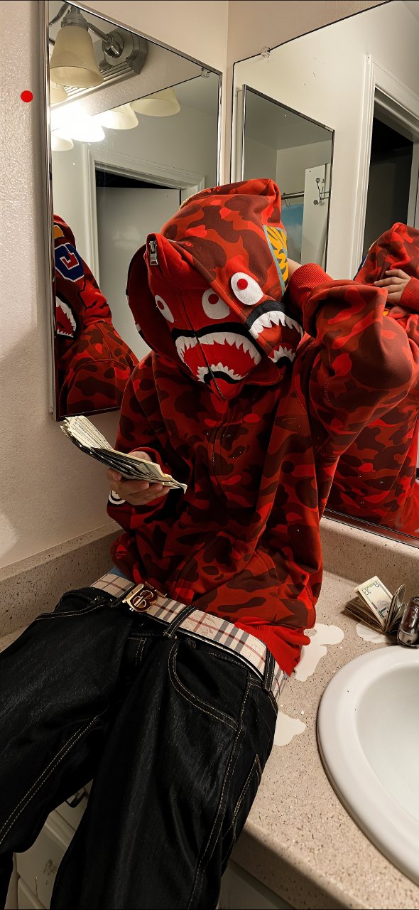 A person in a red camouflage shark-face hoodie takes a bathroom mirror selfie while holding a fan of cards, with dark pants in the foreground.