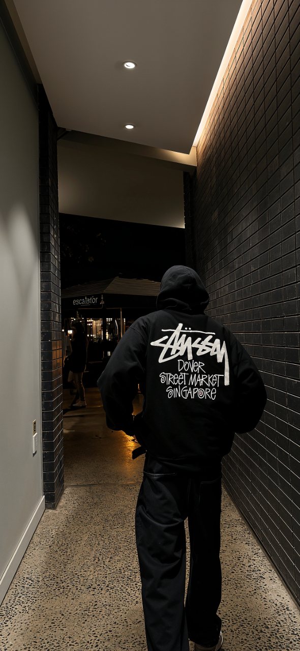 Person wearing a black Stüssy hoodie with Dover Street Market Singapore lettering walking through a dark tiled corridor.
